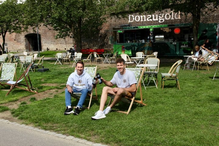 Bei strahlendem Sonnenschein kühle Getränke und Snacks in Liegestühlen oder an kleinen Tischen genießen – für angenehmen Schatten sorgen der alte Baumbestand und weiße Sonnenschirme. Im Hintergrund ragen die historischen Gebäude der Ulmer Altstadt und die Stadtmauer empor: Am 1. Mai 2025 hat die Bar Donaugold eröffnet.